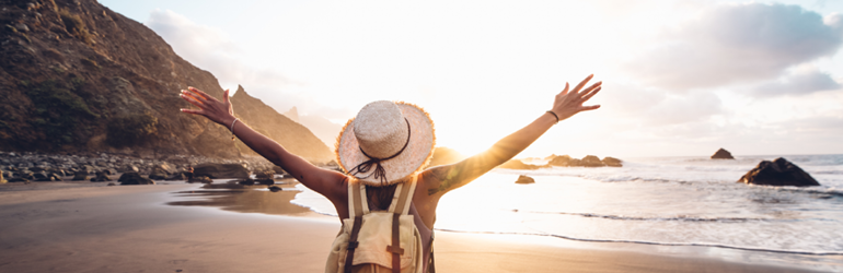 female presenting person wearing a straw hat and yellow shirt stands on beach with arms wide open 