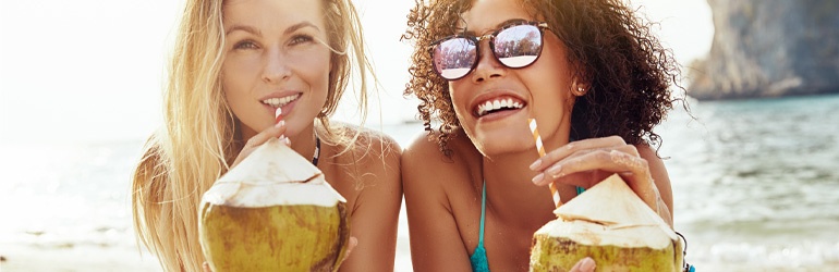 Two women on a beach smile as they sip milk from coconuts through striped straws