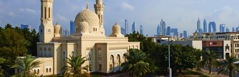 The Jumeirah Mosque surrounded by trees with the Dubai cityscape in the background.