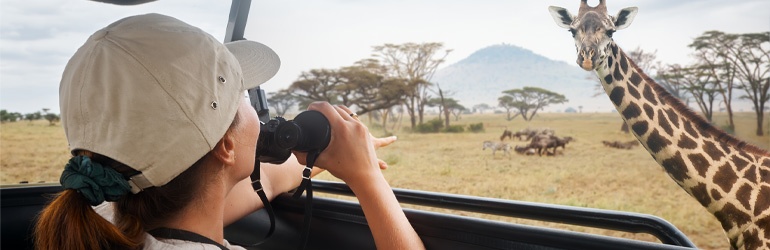 A woman with binoculars watches a giraffe from a safari vehicle
