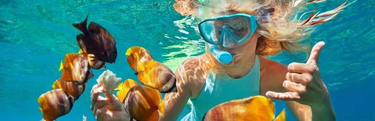 An underwater shot of a woman in a mask and snorkel feeds yellow tropical fish and gives a thumbs-up