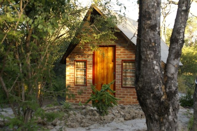 A small brick cabin with a steep roof sits in sand among trees